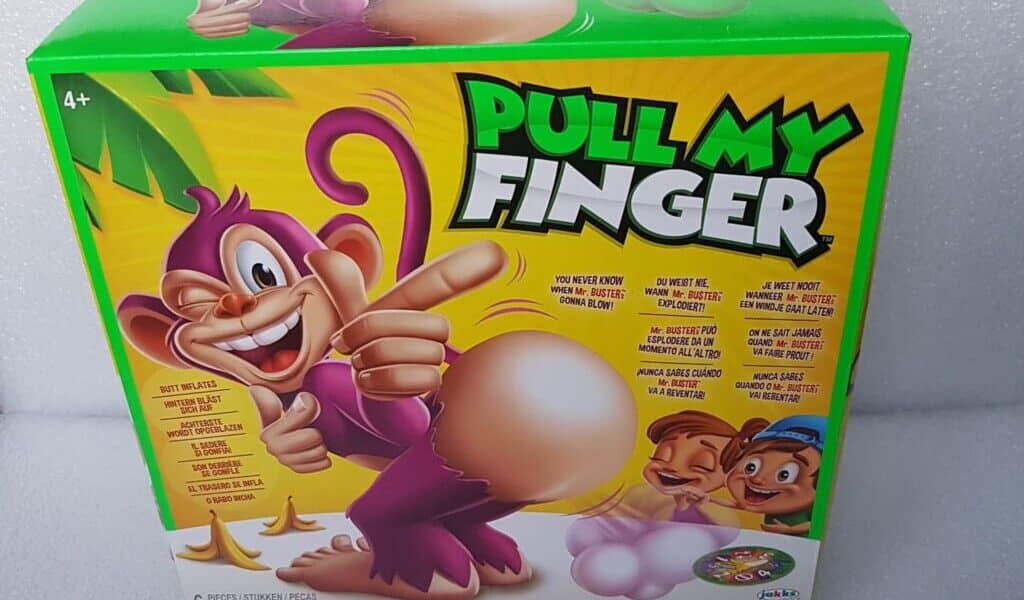 A colourful game box titled "Pull My Finger" features a cartoon monkey pointing to its large, inflated rear. The box shows excited cartoon characters, game instructions, and playful text on a green and yellow background. The game is for ages 4 and up.