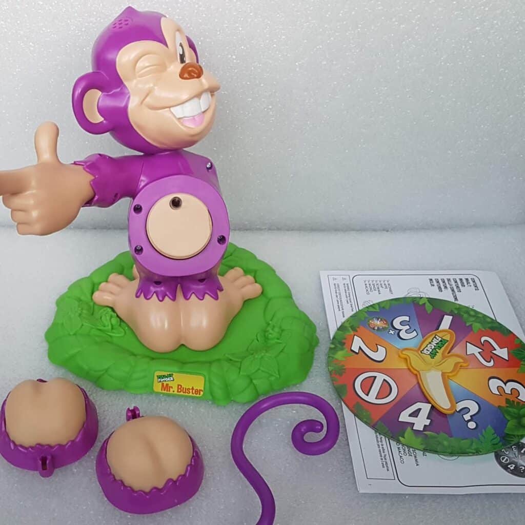 A purple plastic monkey toy with a missing belly and tail is on a green base. Nearby are detachable monkey parts, a colourful spinner with numbers and symbols, and an instruction sheet. The toy is labelled “Mr. Buster.”.