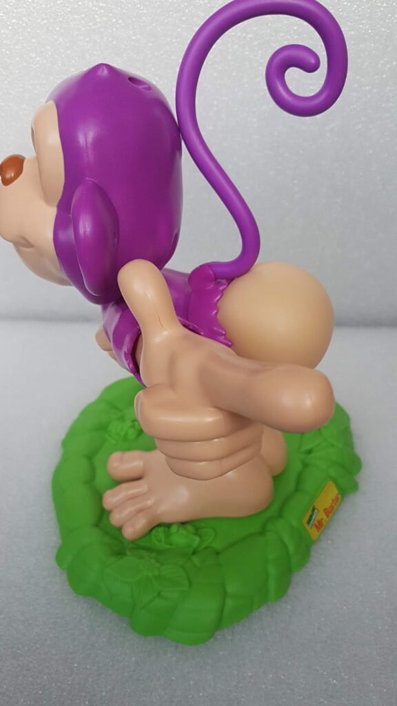 A plastic monkey toy wearing a purple suit stands on a textured green base. The monkey has a curled purple tail and is posed with its large hand pointing forward, while its exposed bottom faces outward. The background is plain and light-coloured.