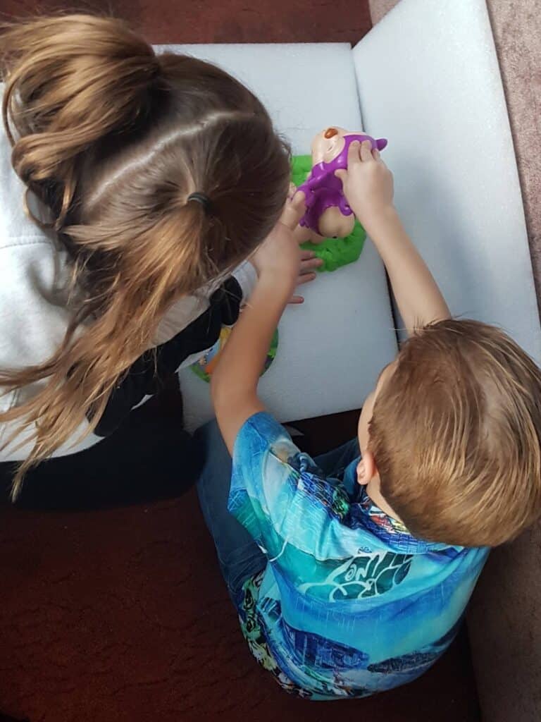 Two children are sitting on the floor and playing with a baby doll. One child holds the doll while the other uses a purple toy medical mask, pretending to give the doll medical treatment. They are sitting close to a white sofa.