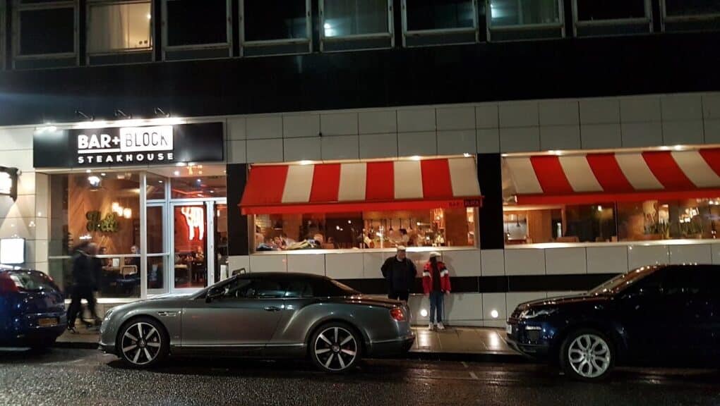 Night-time street view of Bar + Block Steakhouse with a bright sign; large windows reveal diners inside. Two people stand on the pavement. Three parked cars, including a silver convertible, line the wet street in front of the restaurant.