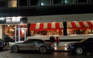 Night-time street view of Bar + Block Steakhouse with a bright sign; large windows reveal diners inside. Two people stand on the pavement. Three parked cars, including a silver convertible, line the wet street in front of the restaurant.