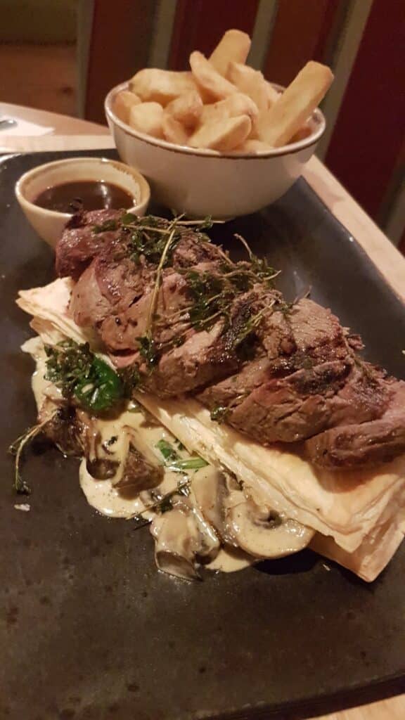 A sliced steak sits atop puff pastry and creamy mushrooms, garnished with herbs, on a dark plate. A small bowl of sauce and a bowl of thick-cut chips are in the background. The meal is served on a wooden table.