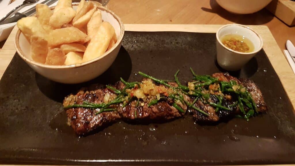 A black rectangular plate holds grilled steak garnished with herbs and a relish, a bowl of thick-cut chips, and a small cup of dipping sauce with visible mustard seeds on a wooden table.