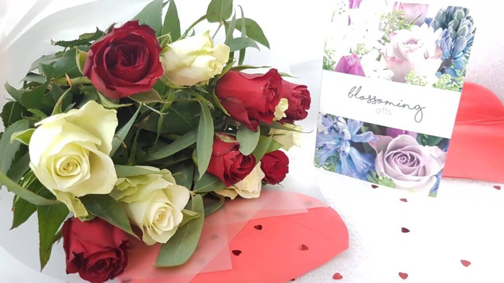 A bouquet of red and white roses with green leaves lies beside a card reading “blossoming gifts,” featuring floral images. The arrangement rests on red paper decorated with small heart-shaped confetti, evoking a romantic, celebratory mood.