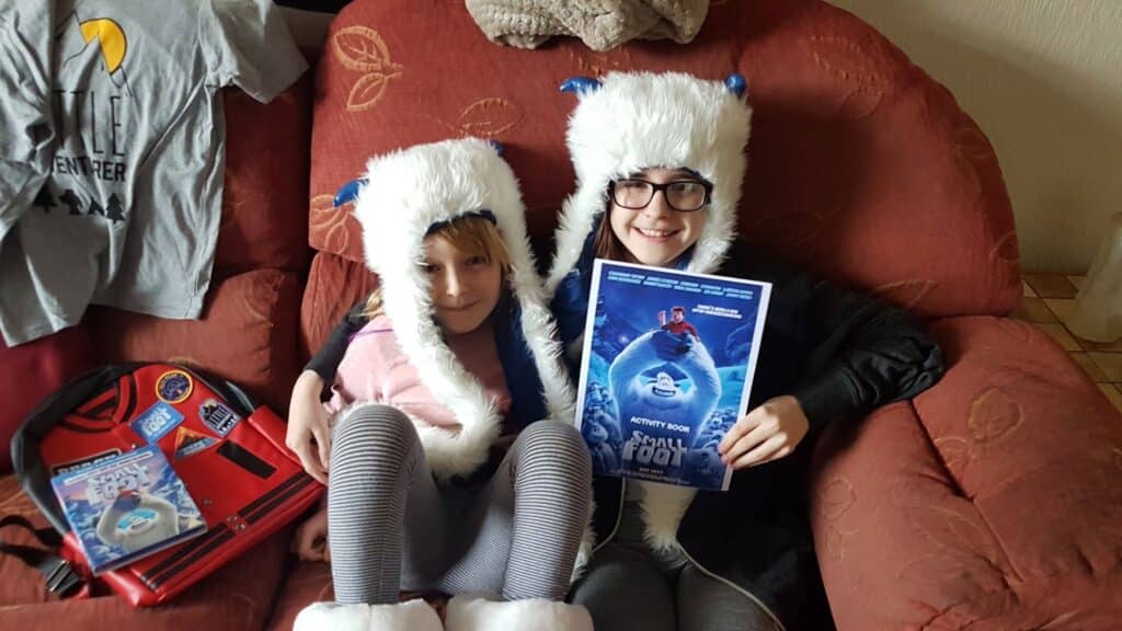Two children sit on a red sofa wearing white fuzzy hats with blue horns and matching white boots. One child holds an "Abominable" film book. A T-shirt, DVD, and rucksack with "Abominable" designs are displayed nearby. Both children are smiling.