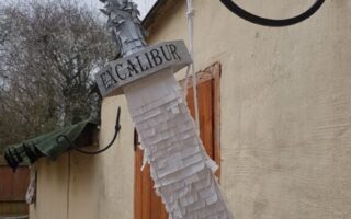 A silver sword prop labelled “Excalibur” hangs from a black metal bracket on a beige stone wall. The sword’s blade is covered with layers of white fringe paper, resembling a piñata. A wooden door and leafless trees are visible in the background.
