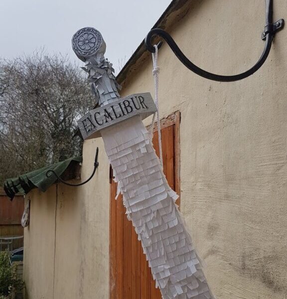 A silver sword prop labelled “Excalibur” hangs from a black metal bracket on a beige stone wall. The sword’s blade is covered with layers of white fringe paper, resembling a piñata. A wooden door and leafless trees are visible in the background.
