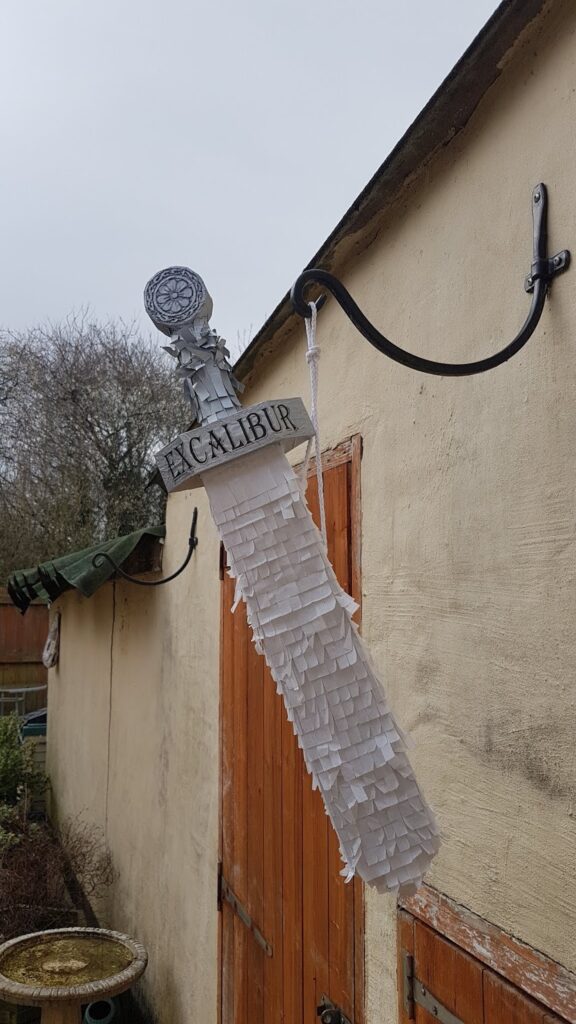 A sword-shaped piñata labelled "EXCALIBUR" hangs from a black metal bracket on an outdoor beige wall. The piñata is covered in white fringe paper, with a detailed silver handle. Wooden doors and bare trees are visible in the background.