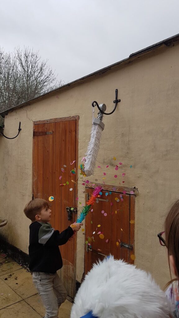 A young boy swings a colourful stick at a white piñata shaped like a mop, hanging outside against a beige wall with a wooden door. Colourful confetti bursts from the piñata. Overcast sky and leafless trees are visible in the background.
