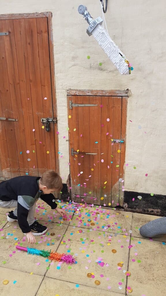 A young child bends down to pick up colourful confetti and sweets from the ground. Above, a silver piñata shaped like a microphone labelled "X-FACTOR" hangs open, with more confetti falling. The scene is outdoors, near two wooden doors on a cream wall.