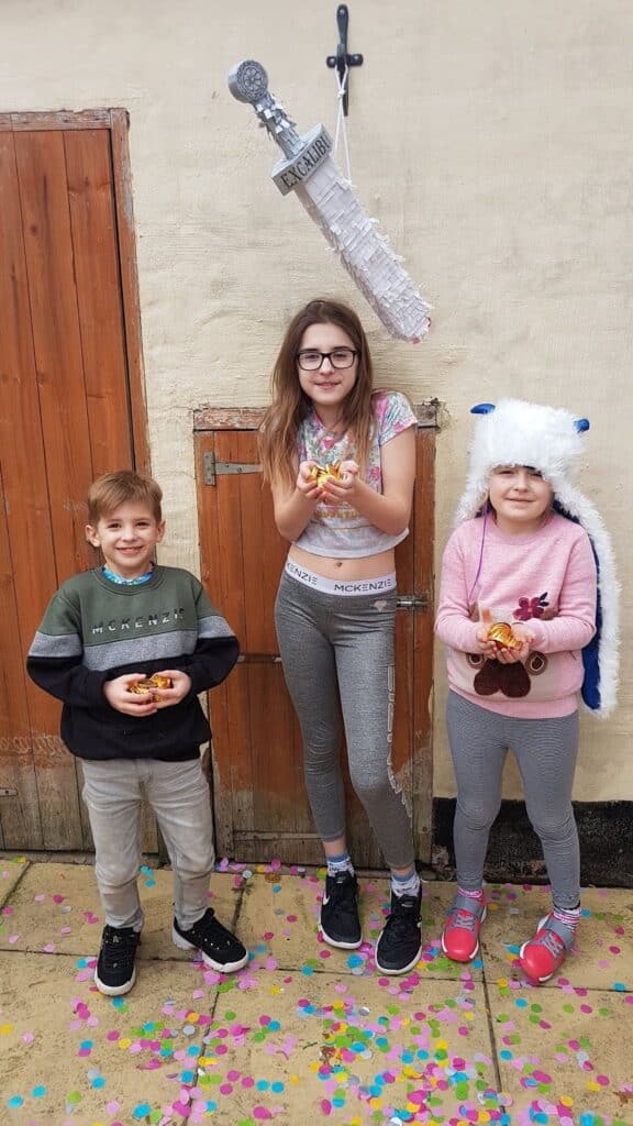 Three children stand outside on a stone patio, smiling and holding sweets or toys with colourful confetti on the ground. Above them hangs a piñata shaped like a microphone labelled "GRAND." The middle girl wears glasses; the right child wears a fluffy white hat.