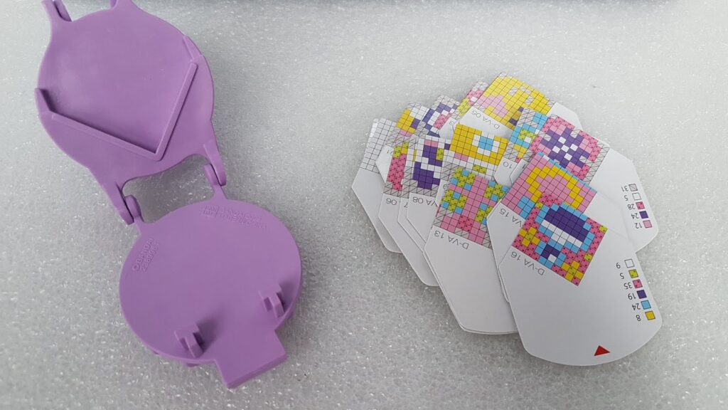 A purple plastic holder is open next to several fan-shaped cards with colourful pixelated grids and small numbers. The items rest on a textured white surface, suggesting a game or puzzle involving colours and patterns.