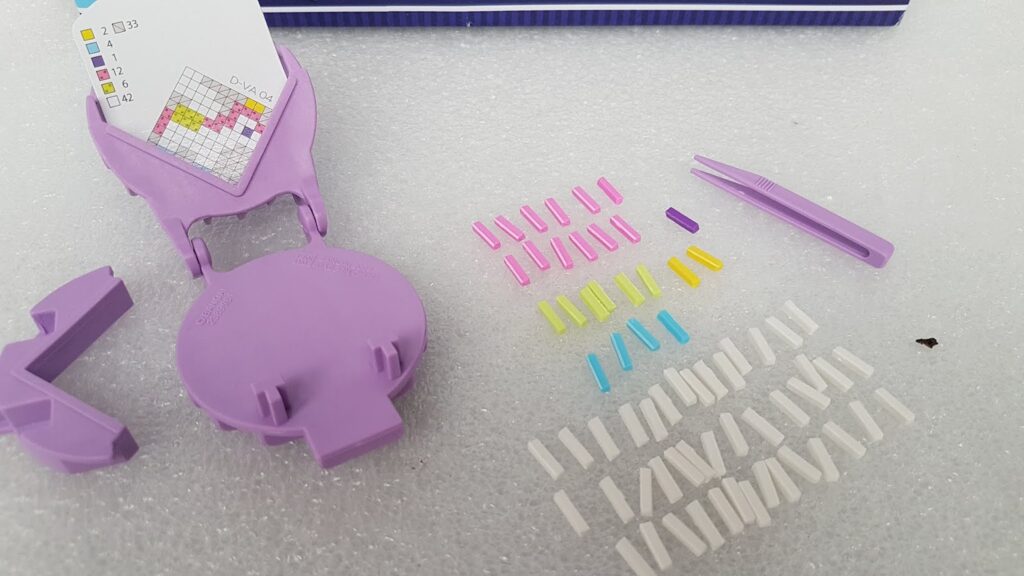 A bead weaving kit is displayed on a white surface. It includes a purple loom, pink tweezers, a colour guide, and small plastic beads sorted by colour: pink, yellow, blue, purple, and white. The items are neatly arranged in distinct groups.