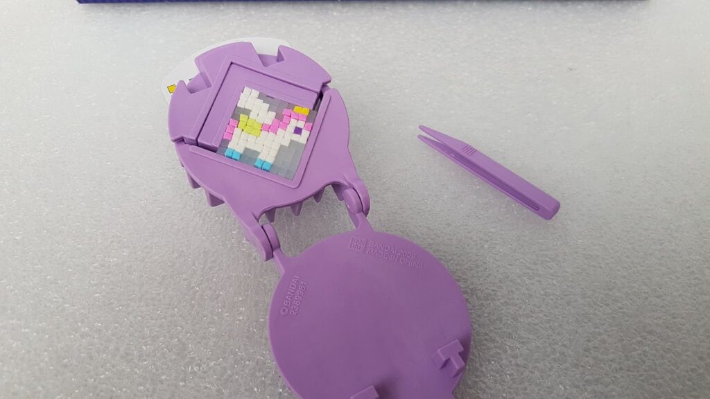 A purple plastic toy with a rectangular grid of small coloured tiles forming a pixelated heart shape, mostly white with pink, yellow, blue, and grey tiles. A matching purple plastic tweezer lies nearby on a textured, light grey surface.