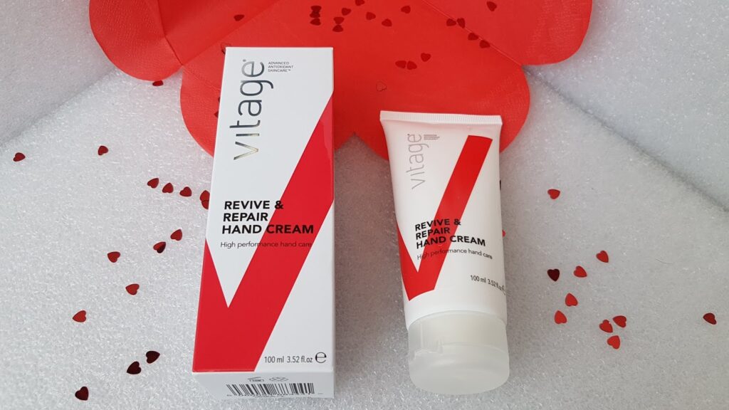 A Vitage Revive & Repair Hand Cream tube and its box are displayed on a white textured surface, surrounded by small red heart confetti. Both the box and tube are white with bold red and black text and a large red "V" graphic.