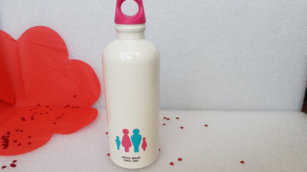A white water bottle with a pink cap stands on a white textured surface. The bottle features a graphic of three human silhouettes in blue and pink. In the background, there is a red heart-shaped fabric and scattered red heart confetti.