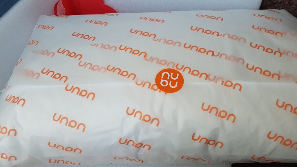 A white parcel wrapped in paper printed with the repeated orange text "unun." In the centre, there is an orange sticker with the white letters "nu" stacked above "ou." The parcel appears to be resting in a white box or container.