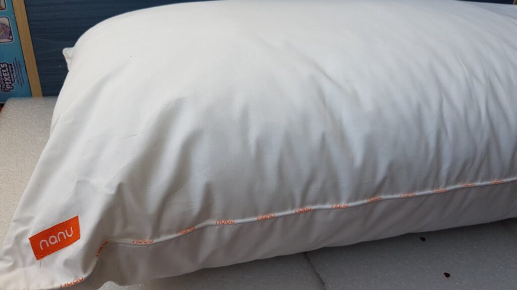 A white pillow with a smooth, plain cover is shown resting on a light surface. An orange fabric tag on the lower left corner displays the brand name "nanu" in white letters. The pillow appears fluffy and well-stuffed.