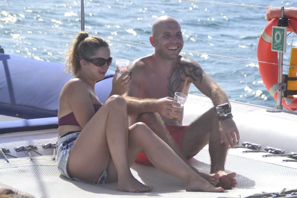 A smiling couple relax on a boat, sitting on a netted deck. The woman wears sunglasses, a burgundy bikini top, and denim shorts while holding a drink. The man, tattooed and shirtless, also holds a drink. The sea glistens in the background.