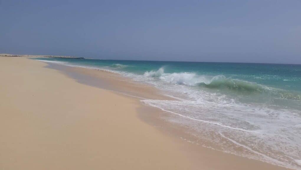 A wide, sandy beach stretches along the left side under a clear, blue sky. Gentle turquoise waves crash onto the shore, creating white foam. The sea meets the sandy beach in a smooth curve, and there are no people or buildings in sight.