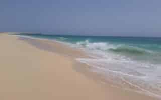 A wide, sandy beach stretches along the left side under a clear, blue sky. Gentle turquoise waves crash onto the shore, creating white foam. The sea meets the sandy beach in a smooth curve, and there are no people or buildings in sight.
