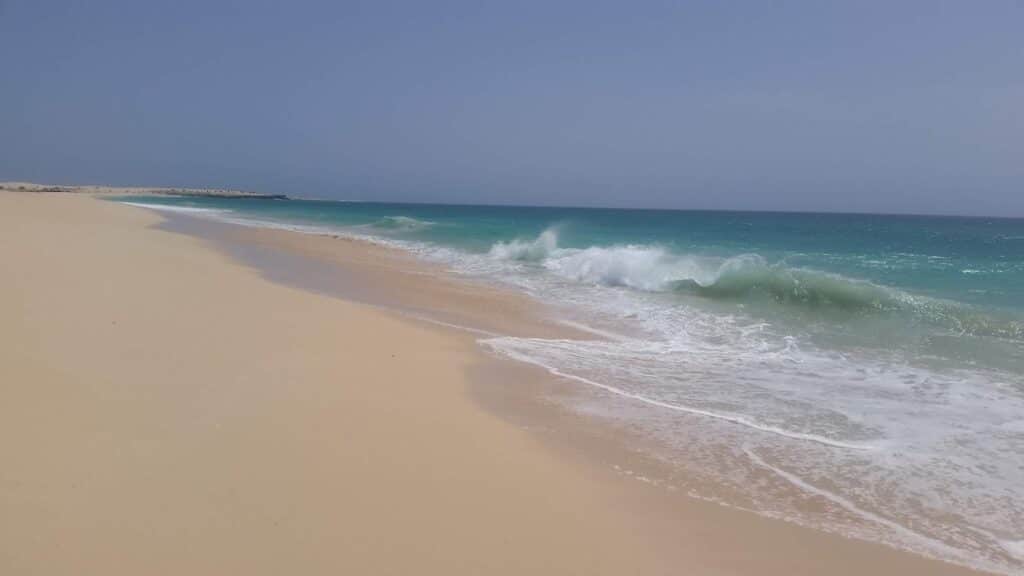 A serene, empty beach with smooth, golden sand stretches into the distance. Gentle turquoise waves crash lightly along the shoreline under a clear blue sky, creating white foam. No people, objects, or buildings are visible, evoking a peaceful, natural atmosphere.