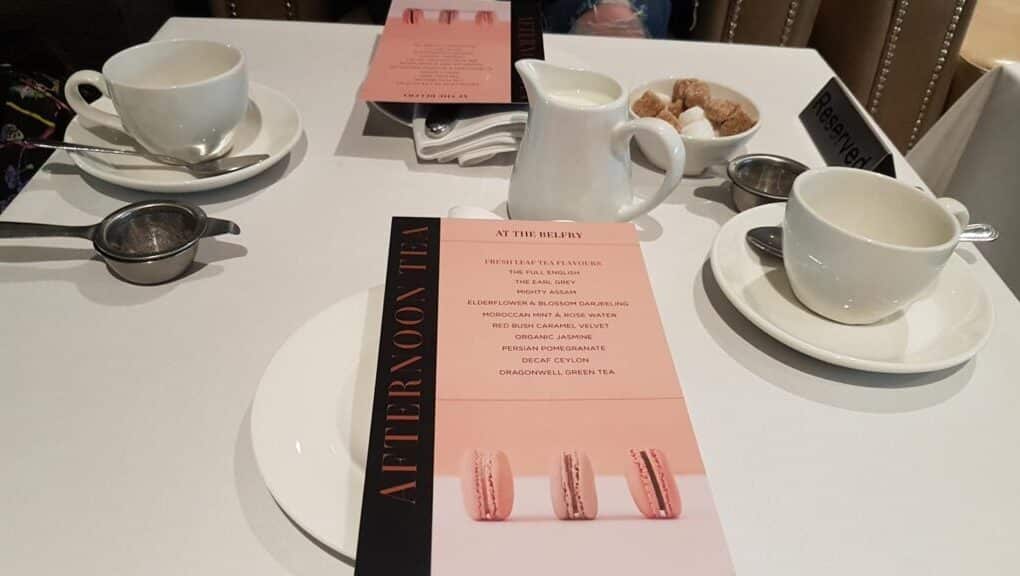 A table set for afternoon tea with two white teacups, metal tea strainers, a milk jug, a bowl of sugar lumps, and menus featuring images of macaroons. The table has a "Reserved" sign and is arranged neatly for two people.
