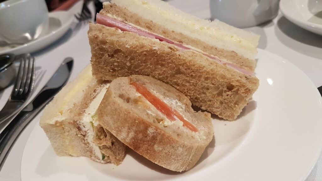 A white plate holds three types of tea sandwiches: one with ham and cheese on wholemeal bread, one with a creamy filling on white bread, and a round sandwich with smoked salmon and cream cheese on a crusty roll. Cups and cutlery sit in the background.