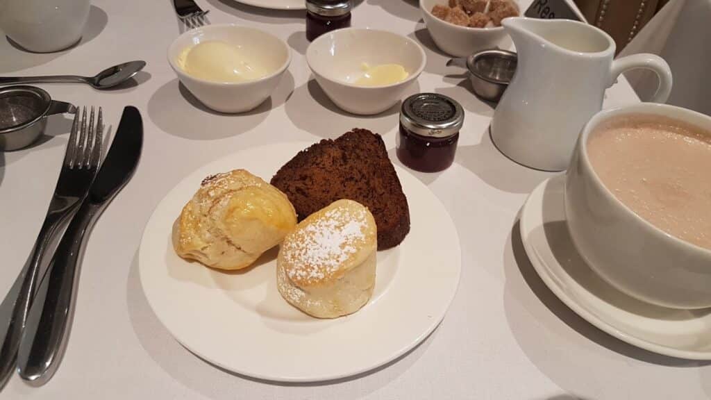 A white plate holds two scones dusted with icing sugar and a slice of dark cake. Surrounding it are bowls of clotted cream, butter, and sugar lumps, a cup of frothy tea, small jam jars, and cutlery on a white tablecloth.