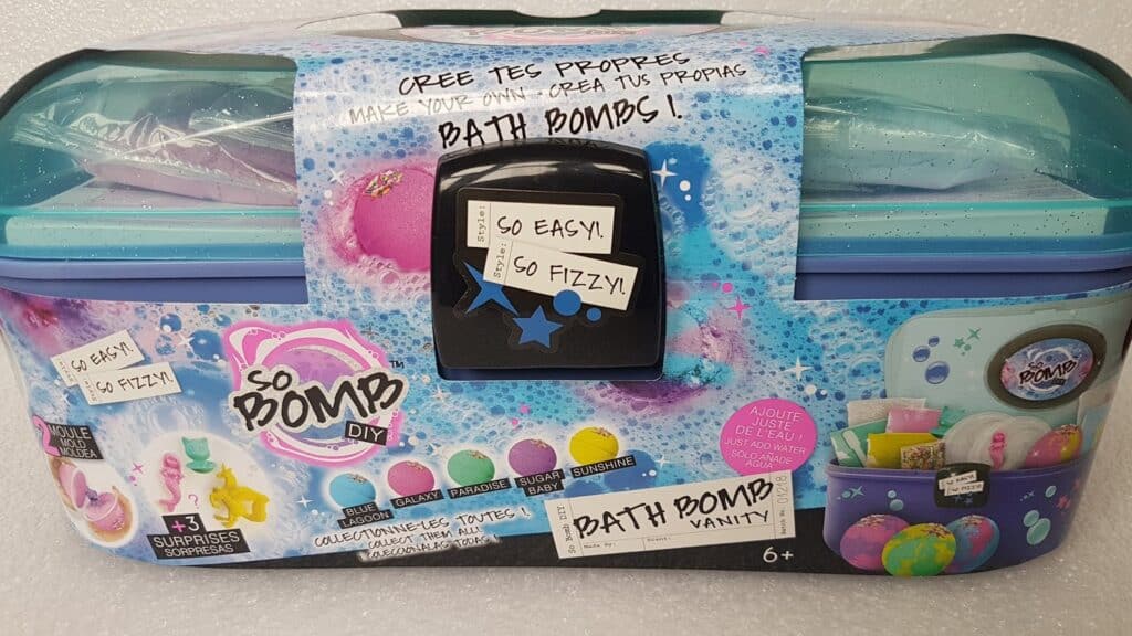 A colourful “So Bomb DIY Bath Bomb Vanity” kit with a teal lid, black latch, and vibrant packaging. Text and images show bath bombs, kit contents, and decorations. Labels read “So easy, so fizzy!” and “3 surprises.” For ages 6+.