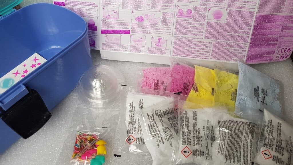 A DIY bath bomb kit is displayed, featuring a blue plastic container, colour powders in pink, yellow, and blue packets, clear packets with white powder, a clear plastic mould, small colourful decorations, and an instruction sheet with text and diagrams.