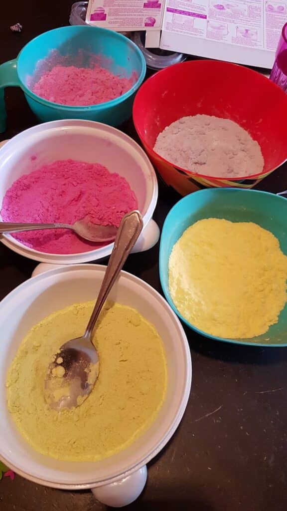 Five bowls, each filled with a different brightly coloured powder (pink, grey, yellow), are arranged on a dark table. Two bowls have metal spoons. An instruction sheet and cup are visible in the background. The scene suggests a craft or baking activity.
