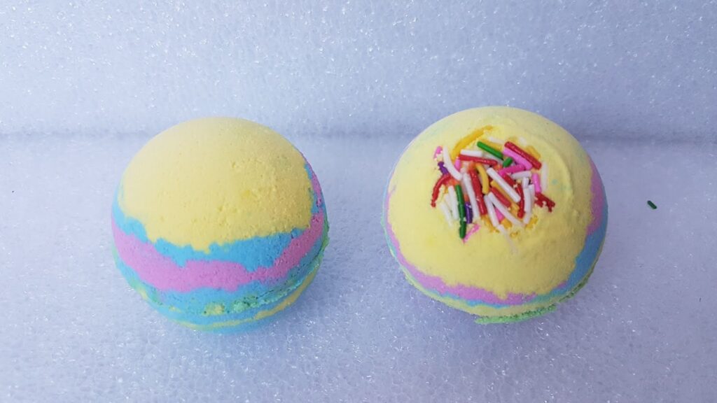 Two round bath bombs rest on a white textured surface. Both are layered with pastel yellow, blue, and pink colours. The right bath bomb is topped with colourful sweet-like sprinkles, while the left one has a smooth top without decorations.