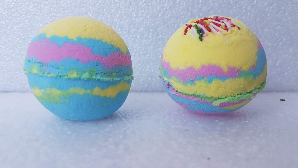 Two round bath bombs with pastel stripes of blue, yellow, pink, and green sit on a white textured surface. The bath bomb on the right has colourful sprinkles embedded on top, while the one on the left has a smooth surface.