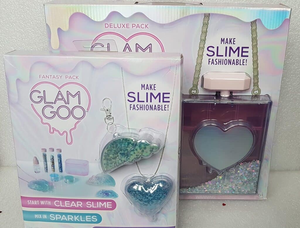 Two "Glam Goo" slime kits are displayed. The front box shows “Fantasy Pack” with clear slime, sparkles, and heart- and crescent-shaped containers. The back box, “Deluxe Pack,” includes a heart-shaped handbag and necklace, advertised to “make slime fashionable!”.