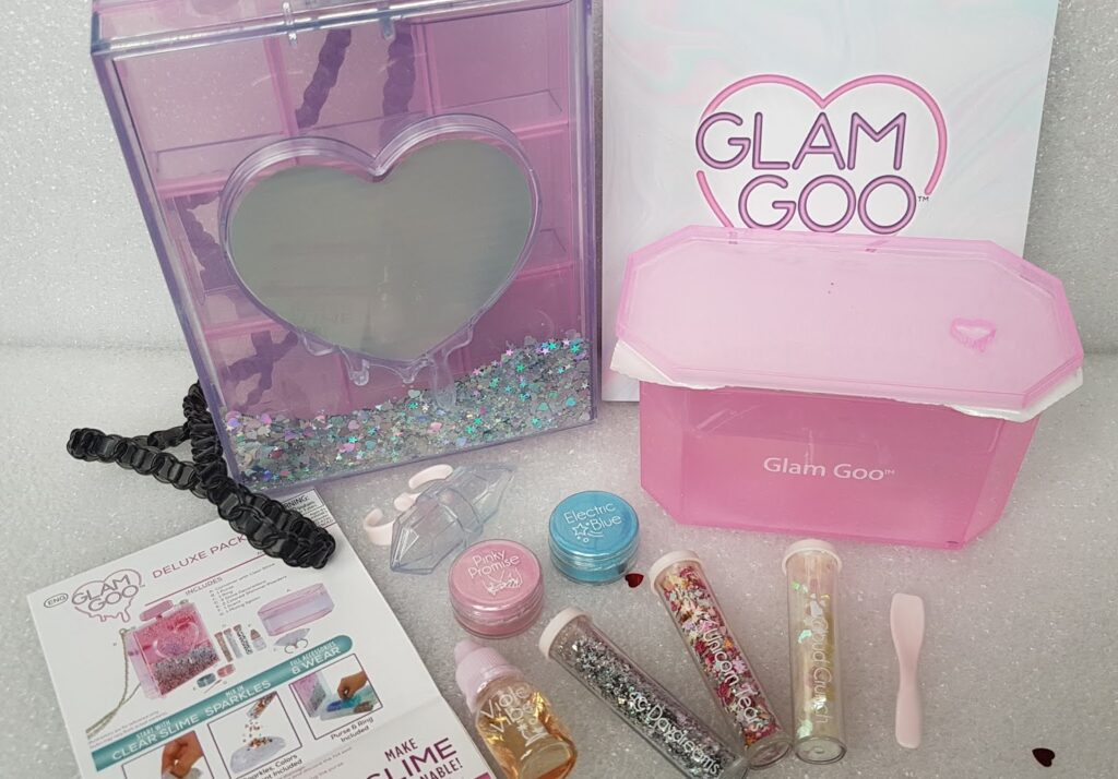 A Glam Goo deluxe slime kit with a heart-shaped mirrored case, pink storage container, glitter, coloured powders, mixing spoon, black scrunchie, rings, clear slime, decorative charms, and an instruction sheet arranged on a white surface.