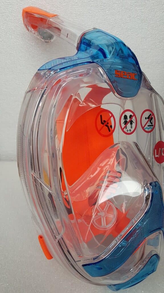 A close-up of a clear, full-face snorkelling mask with orange and blue accents. The brand "SEAC" is visible on the top. Safety warning icons are displayed, including no diving and adult supervision required. The mask rests against a white textured background.
