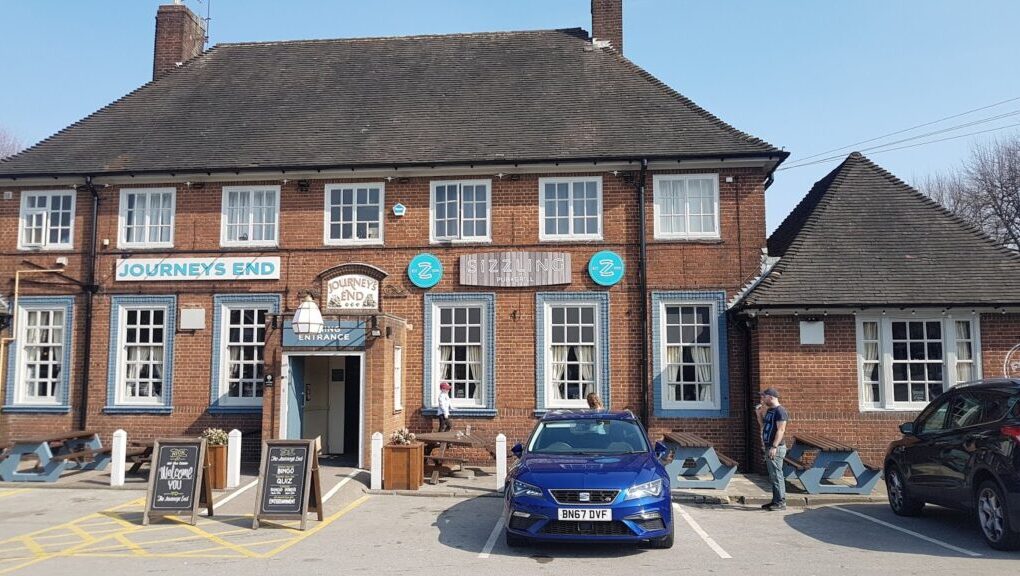 A red-brick building labelled “Journey’s End” with blue-trimmed windows and doors. A blue car is parked in front. Two people stand outside—one near the entrance, another by the car. Outdoor seating and menu boards are visible. It is a sunny day.