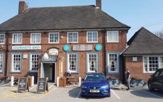 A red-brick building labelled “Journey’s End” with blue-trimmed windows and doors. A blue car is parked in front. Two people stand outside—one near the entrance, another by the car. Outdoor seating and menu boards are visible. It is a sunny day.