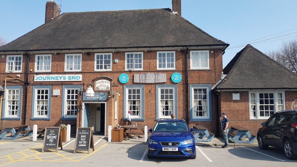 A red-brick building labelled “Journey’s End” with blue-trimmed windows and doors. A blue car is parked in front. Two people stand outside—one near the entrance, another by the car. Outdoor seating and menu boards are visible. It is a sunny day.