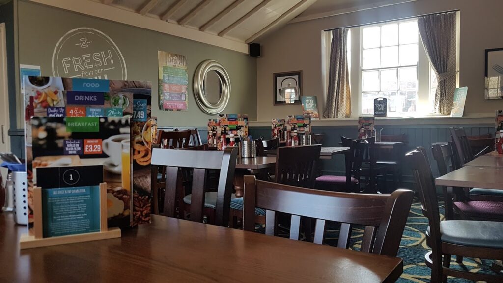 A cosy, well-lit restaurant with wooden tables and chairs, colourful menu stands on each table, a large window with curtains letting in daylight, wall decor including a round mirror and poster, and blue-green walls with “FRESH” painted on one.