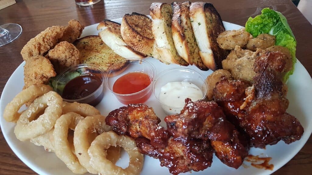 A white plate holds fried chicken goujons, onion rings, barbecue chicken wings, breaded mushrooms, slices of toasted garlic bread, lettuce, and three dipping sauces—barbecue, ranch, and sweet chilli—on a wooden table.