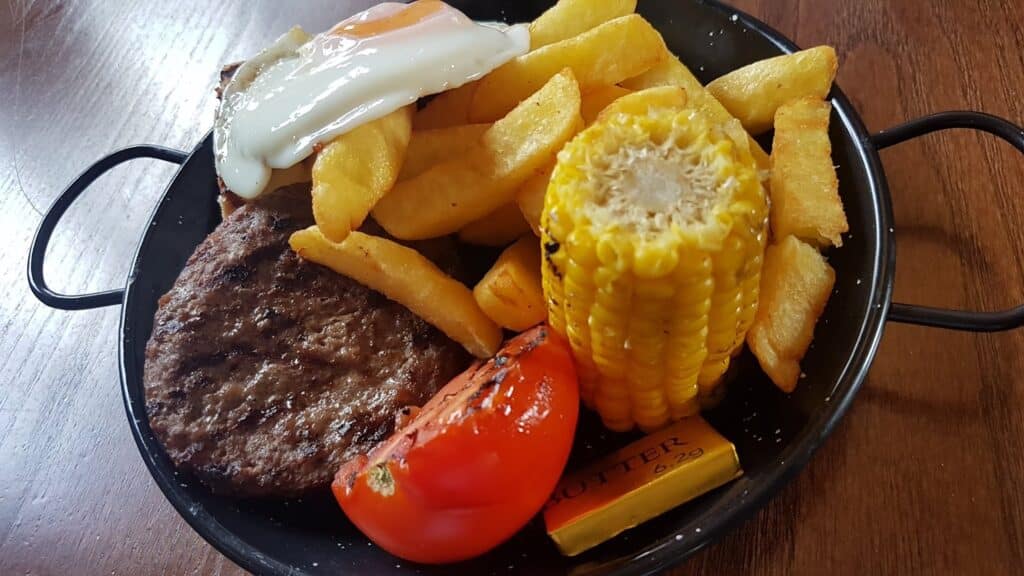 A black round pan holds a grilled beef burger, thick-cut chips, half a grilled tomato, corn on the cob, a pat of butter, and a fried egg with a runny yolk, all set on a brown wooden table.