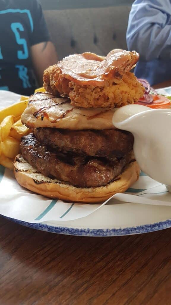 A large burger with two beef patties, grilled chicken, a fried onion ring, and crispy bacon sits on a bap. It is topped with a generous pour of white sauce, accompanied by golden chips on a plate. Two people are partly visible in the background.