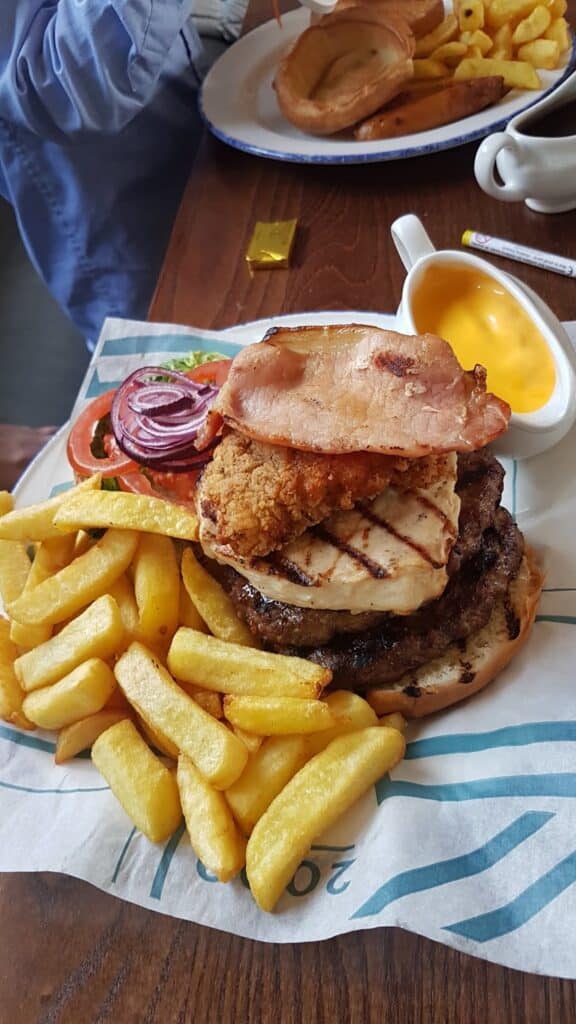 A burger topped with grilled chicken, bacon, fried chicken, tomato, lettuce, and red onion sits on a paper-lined tray, served with thick-cut chips. A small dish of yellow cheese sauce and another plate of food are visible in the background.