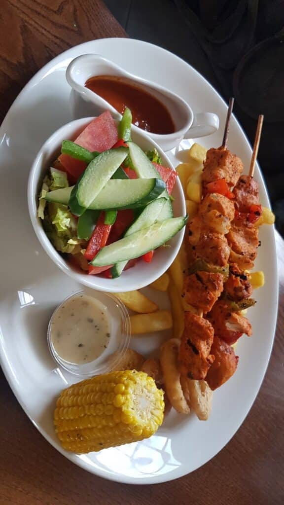 A white plate holds two grilled chicken kebabs, chips, corn on the cob, a small bowl of green salad with cucumbers and tomatoes, a small pot of creamy dipping sauce, and a gravy boat with reddish-brown sauce.