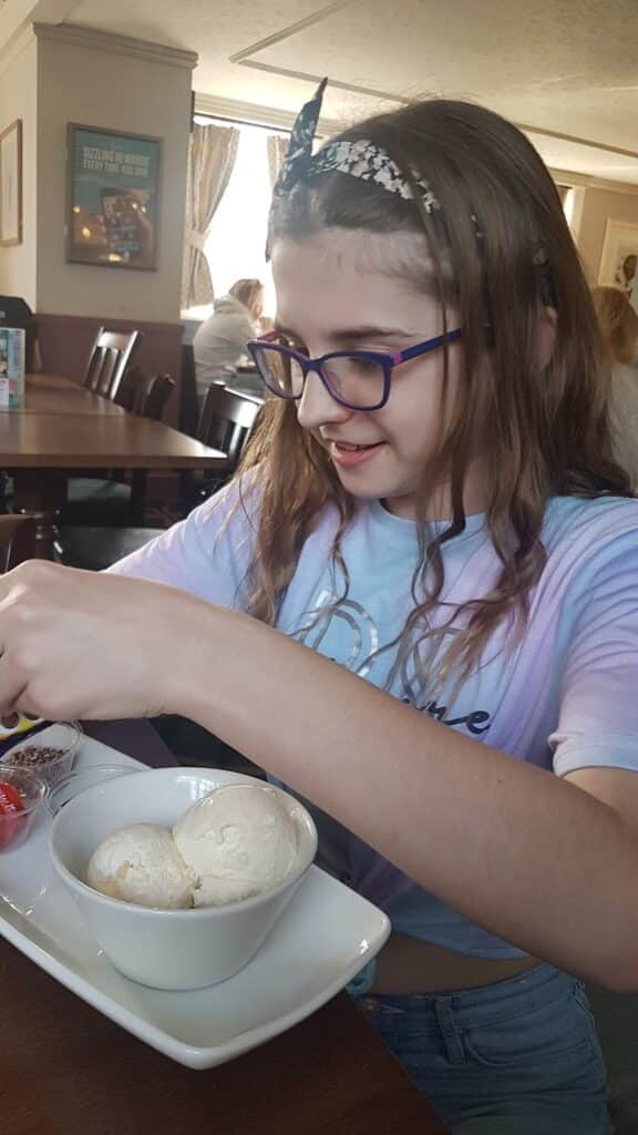 A smiling girl with long brown hair, a hairband, and purple glasses sits at a table in a restaurant, looking down as she prepares to add toppings to a bowl of two scoops of vanilla ice cream in front of her. Sunlight streams in from a window behind her.