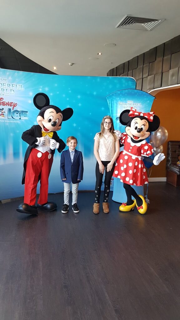 Two children, a boy in a suit and a girl in casual clothes, stand indoors between people dressed as Mickey Mouse and Minnie Mouse. They are smiling in front of a blue backdrop with Disney On Ice branding. The setting looks festive and cheerful.