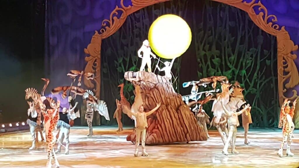 Performers in animal costumes, including lions, zebras, and giraffes, stand on a set resembling Pride Rock from "The Lion King." A figure dressed as Rafiki holds up a small lion cub in front of a glowing yellow sun, recreating the iconic scene.